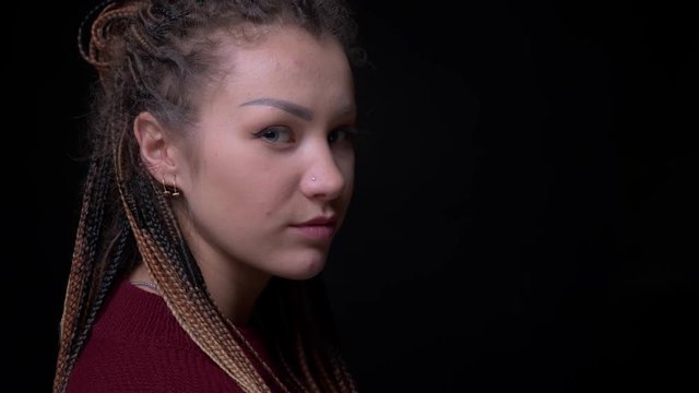 Young cool brunette girl with piercing and dreadlocks turns to camera and watches calmly into it on black background.