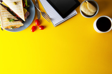 Snacks, fast food concept. Eatting at work place. Fresh club sandwich, vegetables, coffee, potato chips, sweet cookies. Tablet screen. Yellow background.