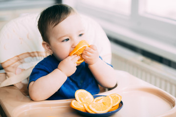 a little girl in a blue t-shirt and a blue plate sitting in a child's chair eating an orange