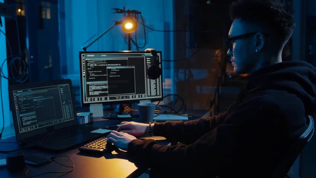 cybercrime, hacking and technology concept - asian male hacker in glasses using computer for cyber attack in dark room at night