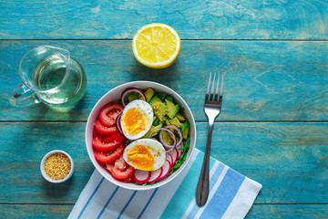 healthy bowl with boiling egg fresh tomatoes avocado radish arugula red onion sesame seeds fork napkin lemon  olive oil on blue wooden table top view copy space
