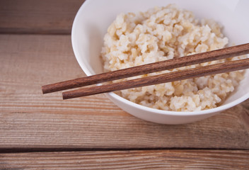 Cooked brown rice in white bowl with chopsticks on the wooden background.