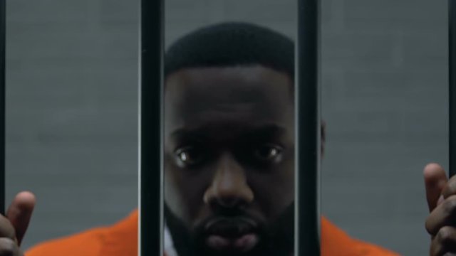 Upset Afro-American prisoner holding bars in cell and looking at camera, guilty
