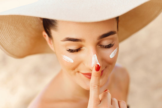 Beautiful Young woman with sun cream on face. Girl holding sunscreen bottle on the beach. Female in hat applying  moisturizing lotion on skin.Skin care. Sun protection. Suntan