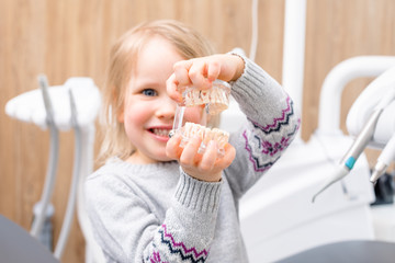 Little girl shows artificial model of child jaw in pediatric dental clinic