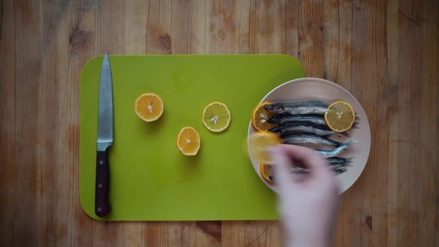 Top view of the process of making a small silver fish, male hands cover capelin with fresh lemon slices, lying in a beige plate on a wooden table.