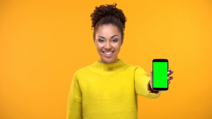 Smiling young lady in yellow sweater showing smartphone with green screen, app