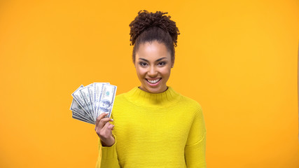 Excited afro-american female showing dollars on bright background, investment