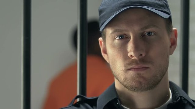 Prison guard looking at camera, maintaining order, black prisoner on background