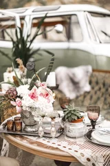 Schöne Gläser, Kerzen, Blumen stehen auf einem fruchtbar dekorierten Tisch im rustikalen Boho-Stil. Ein großes grünes Sofa und ein Hippie-Bus im Hintergrund. Sommerliche Wüstenatmosphäre. Hochzeitsfeier © sofiko14