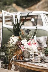 Beautiful glasses, candles, flowers stand on a fruitful decorated table in rustic boho style. A large green sofa and hippy bus on the background. Summer desert atmosphere. Wedding party