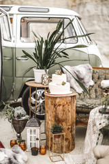 Stylish rustic decor composition of the vintage sofa, golden iron decorations with cactus succulents and plants, candles and wedding cake on wooden log. Rustic wedding decor and hippy bus