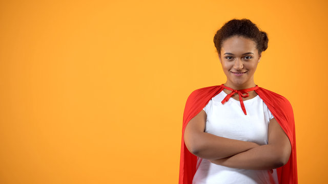 Confident young lady in red cape on bright background, powerful wonder woman