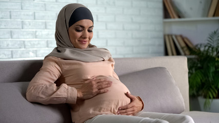 Smiling pregnant woman holding belly, resting on home sofa, maternity happiness