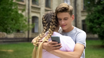 Teenager using cell phone while hugging girlfriend, absorbed by social networks