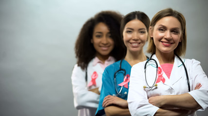 Confident multiethnic nurses with pink ribbons, international breast cancer sign