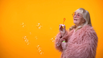 Positive aged woman in funny pink coat and round sunglasses making soap bubbles - Powered by Adobe