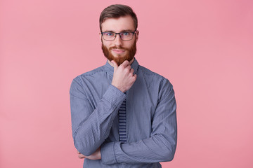 A confident young bearded man in glasses, holds his hand on chin, thinks about something, build a...