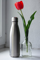 Thermo water bottle of steel near red tulip flower in water, on window background.