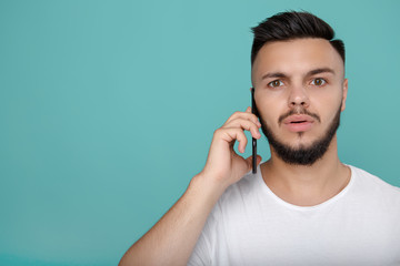 Handsome young bearded hipster in white t-shirt using phone and talking with bright smile. Copy space.