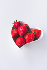 Top and close up view of fresh red strawberries in a white heart shaped porcelain bowl isolated on white background. Love healthy food concept