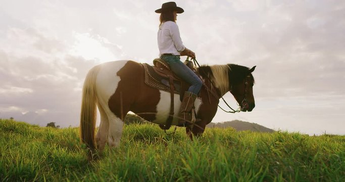Cowgirl sitting on horse in green fields at sunset, slow motion orbit shot of horseback rider