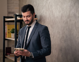 Good looking handsome businessman in the office standing by his desk and counting money. wearing suit and a tie