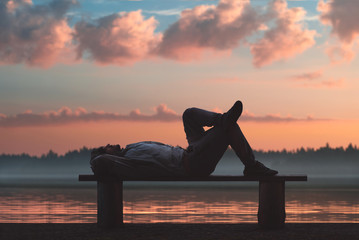 A man is lying on a wooden bench in a picturesque place. Lake and forest in the background.