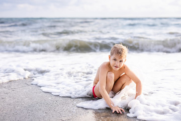 Fototapeta na wymiar Little cute caucasian toddler boy with blond hair plays at the beach in sea waves on sunny summer day. Family vacation with kids fun happiness closeness unity with nature. Authentic lifestyle