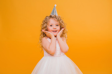 Young blond girl in birthday party princess hat hands spread up screaming isolated on a yellow background,Young beautiful girl wearing birthday cap over isolated background smiling with happy face