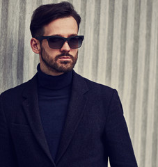 Fototapeta premium Fashion beard style business handsome male model in fashion sunglasses and blue coat with serious concentrated look posing in style clothing on wall outdoors background. Closeup