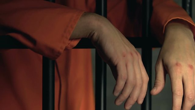 Criminal with beaten hands standing behind prison bars, conflicts in jail