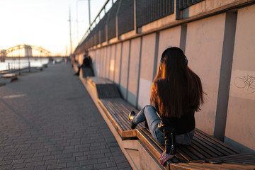 Fototapeta na wymiar Young woman listens to music in closed headphones through her phone wearing a leather jacket and jeans at a sunset near river Daugava