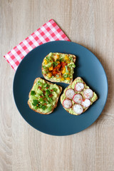 Three slices of toast with mashed avocado and various vegetable and herb toppings. Top view.