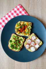 Three slices of toast with mashed avocado and various vegetable and herb toppings. Top view.