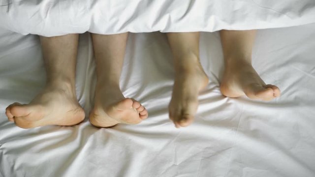 Feet dancing under a white blanket, on a white bed, top view.4k