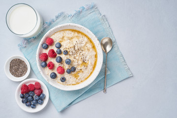 Oatmeal with berries, chia, maple syrup and glass of milk on blue light background. Top view, copy space. Healthy diet breakfast