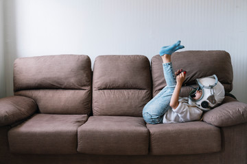 Adorable little girl sitting on couch. He is lying on couch while playing with a video game console at home