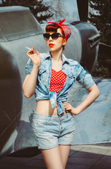 Pin up beautiful girl with red hair stands against the background of an old plane