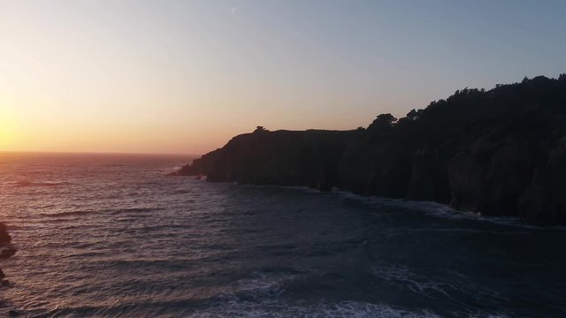 Drone captures a setting sun at golden hour over a lonely cove with a rocky beach. Haze and setting sun create a dreamy and surreal vibe.