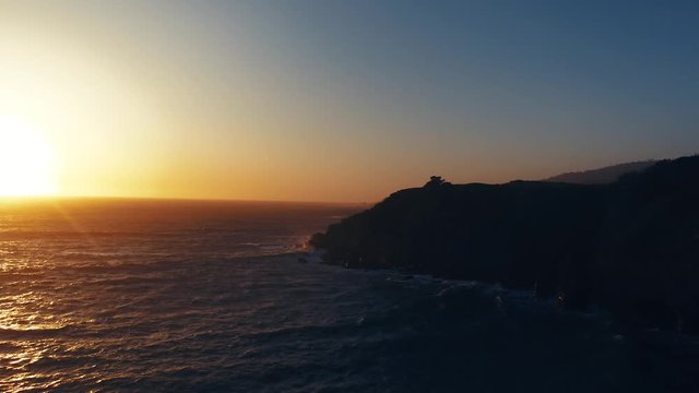Drone captures a setting sun at golden hour over a lonely cove with a rocky beach. Haze and setting sun create a dreamy and surreal vibe.