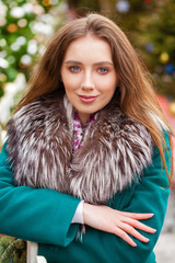 Happy young brunette woman in a green coat with a fur collar, winter park outdoors