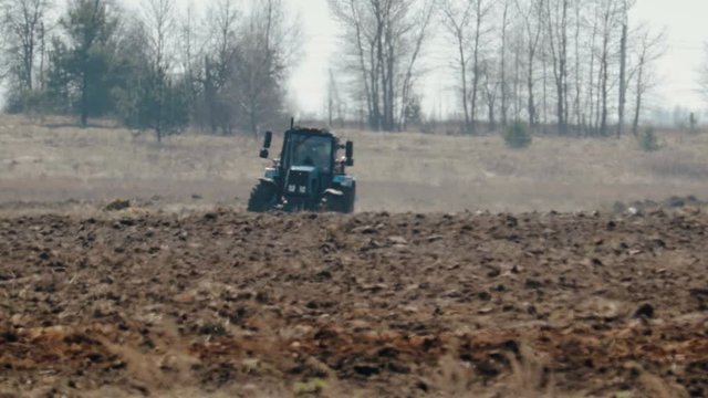 Static shot of a blue tractor with big black wheels and a powerful grouser, heavy plowing the dark fertile soil. The warmth from the ground and the motor distorts the image