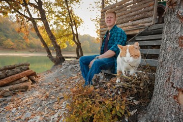 Blurred redhead man in a plaid shirt and purple t-shirt is sitting on the steps of the stairs of a wooden house on the lake in the autumn forest. In the foreground in focus is a white-red cat.