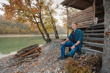 Redhead man in a plaid shirt and purple t-shirt is sitting on the steps of the stairs of a wooden house on the lake in the autumn forest. White-red cat is on the doorstep.