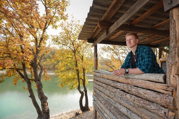 Fototapeta premium Redhead man in a plaid shirt and purple t-shirt looks into the distance from a wooden gazebo on the lake in the autumn forest.