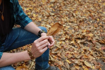 Man in a plaid shirt and purple t-shirt  holds in his hand a fallen and dead leaf in the autumn forest. Autumn, walking and wilting nature concept.