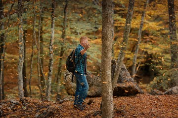 Naklejka premium Redhead man in a plaid shirt and purple t-shirt walks through the autumn forest with his hands raised and enjoys the leaves falling from the trees. Autumn, travel and happiness concept.