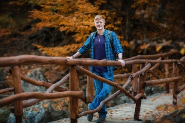 Redhead man in a plaid shirt and purple t-shirt stands on a wooden bridge over a mountain stream in the autumn forest in the Grand Canyon of Crimea. Autumn, travel and tourism concept.