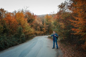 Redhead man in a plaid shirt and purple t-shirt with backpack hitchhikes on the road in the middle of the autumn forest. Autumn, travel and tourism concept.